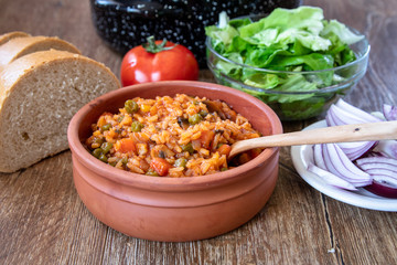 Fried rice with vegetables and spices in a vintage clay bowl and tomato, lettuce, onion and bread on wooden table
