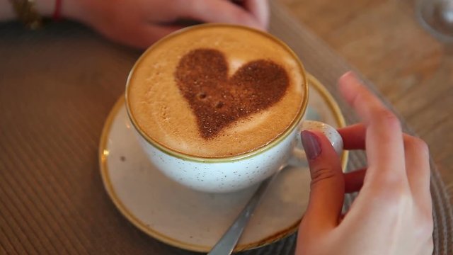 A girl sits at a table in a cafe with a mug of coffee with a painted heart.