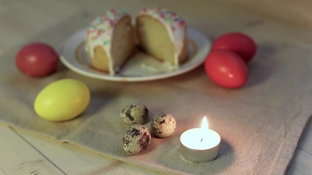Happy Easter concept, background for a greeting card: closeup burning light candle, colorful Easter eggs in the background and Easter cake cut in two pieces