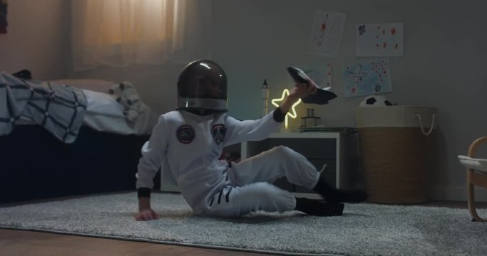 Cute little child girl wearing astronaut suit playing with toy shuttle space ship rocket at home, bedroom interior evening shot. 4K UHD RAW FOOTAGE