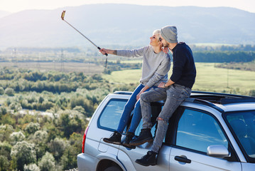 Young hipster couple in love making selfie using smart phone while sitting on the roof of off-road car on mountain background.