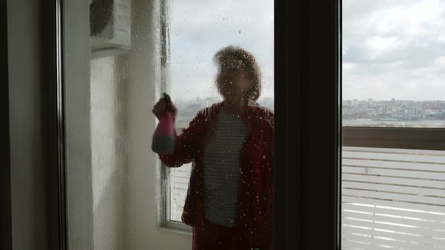 SIDE VIEW: Mature woman sprays window cleaner on large window and washes it indoors