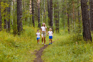 a Caucasian slender smiling woman and two pre-school children in white t-shirts run along the path, holding hands in the summer outdoors