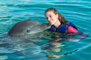 Fototapeta premium Dolphin kiss young woman in blue water. Smiling woman swimming with dolphin. Blue ocean water background.