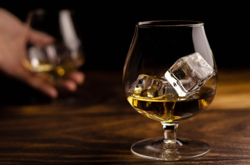 Whiskey / cognac glass with ice in a hand on a wooden background. Dark backdrop.