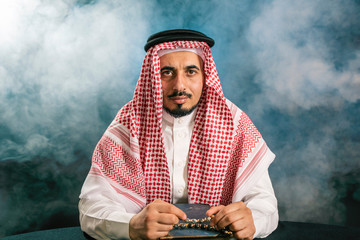 Oriental man in a white, traditional, national, Arabic dress dishdash, sitting in a haze with a rosary and the old Quran.