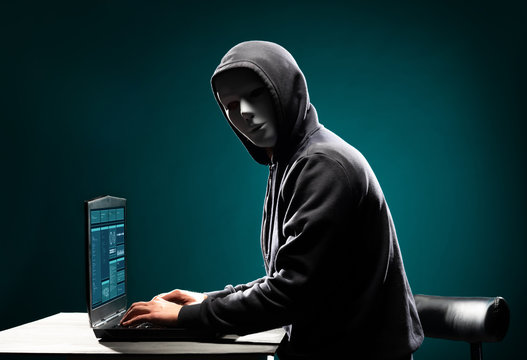 Computer hacker in white mask and hoodie. Obscured dark face. Data thief, internet fraud, darknet and cyber security concept.