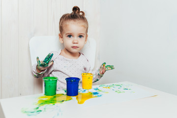 Beautiful little girl draws with finger paints on a white sheet of paper. Creative child development in kindergarten or free time at home