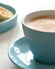 Close up of cup of frothy white coffee with plate of muffins in background