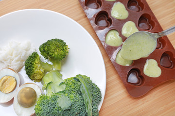 Raw of baby food (broccoli, egg and rice) in white plate and mashed baby food in container for freezing on wooden board.