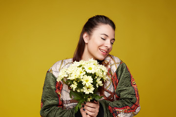 Jubilant upbeat mixed raced woman with broad shining smile, keeps eyes shut, bites tongue, wears colorful hoodie, holds fresh flowers with pleasant scent, models over yellow studio wall background.