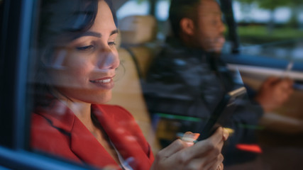 Young Woman and Man Riding on the Backseat of a Car in the Evening. They Both Use Smartphones, Browse in Internet and Do Business Work. Camera Shot made from Outside the Car.