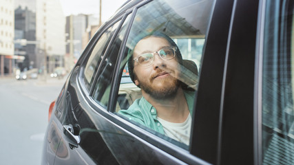 Stylish Young Man Rides on a Passenger Back Seat of a Car, Looks out of the Window in Wonder. Big City View Reflects in the Window. Shot Made Outside the Car. 