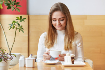 Attractive model sits in chair and looks in cup of coffee. Dreaming girl with blond hair wears white dress, holds cup in two hands, looks delighted, enjoys pleasant atmosphere of cosy cafe.