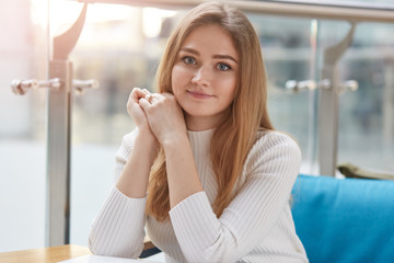 Portrait of attractive young girl being in good mood, has rest in cafeteria after lectures in university, sits on comfortable blue sofa, wears casually, looks at camera, keeps hands folded under chin.