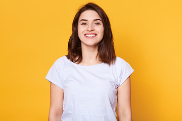 Obraz premium Portrait of emotive good looking caucasian woman laughs while looking directly at camera and standing against yellow background. Charming brunette female with toothy smile looks happy, has great news.