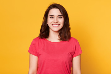 Obraz premium Portrait of young beautiful girl has perfect skin and tooyht smile, stands smiling, looking directly at camera, has happy facial expression, looks satisfied, posing isolated over yellow background.