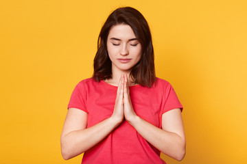 Beautiful young brunette female makes praying gesture, keeps palms pressed together, feels guilty, dressed in casual redt shirt, asks for apologize, isolated over yellow background. People concept.