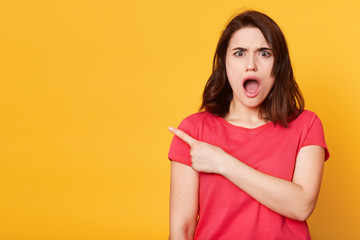 Portrait of astonished woman stands isolated on yellow studio background. Female wears red t shirt,...