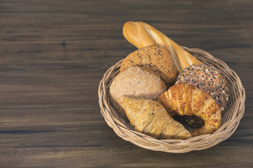 Close up image of variety freshly homemade breads, bun, whole wheat bread with sesame seeds and croissant in wicker basket on the dark wooden table top background