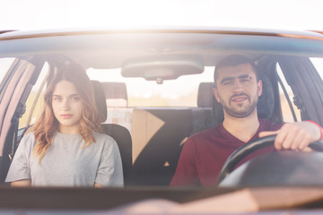 Young man and woman sitting in car, look directly at camera, dressed casually, couple have long...
