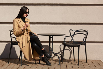 Fototapeta na wymiar Brunette beautiful young lady sits on terrace, drinks aromatic hot coffee, having beige and black look. Attractive woman stops to have good rest before long travel and enjoy warm sunny weather.