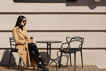 Take away coffee in women's hands. Beautiful young brunette woman wearing brown coat sitting at table in street outdoor cafe and drinking hot beverage, enjoys sunny autumn days, looks thougtful.