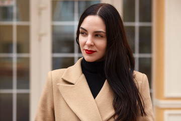 Close up outdoor portrait of young beautiful stylish girl in beige coat and black sweater, has long dark hair, light makeup with red lips, posing near window of cafe, looks thoughtfuly aside.
