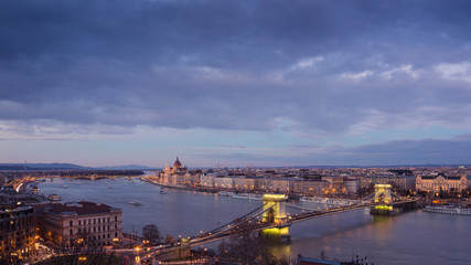 Cityscape of Budapest with bright parliament illuminated by last sunshine before sundown and Danube river with bridge. Pink and purple colors of sky reflecting in water during sunset.