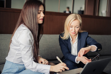 Blonde woman talking with her brunette business partner discussing data on laptop computer, two women entrepreneurs sitting on grey couch in business centre.