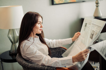 Photo of smiling brown-haired charming woman sitting in arm chair with opened newspaper in hotel room. Business, people and mass media concept