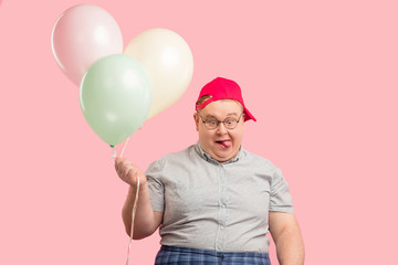 Portrait of plump children animator with comic crazy face expression wearing red baseball cap, holding colorful air balloons, looks at camera like a ninny. People, celebration concept