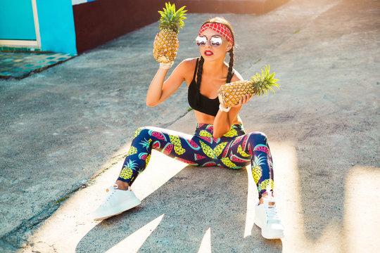 Sports cheerful girl in sports clothes, leggings, top, headband, holding two pineapples and having fun, 80s 90s style