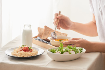 Cropped shot of male having breakfast with salad, pumpkin puree, spaghetti and milk at table in kitchen at home