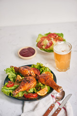 Grilled chicken legs roasted on the grill on dark plate with tomato sauce in a bowl and lettuce leaves, glass mug of beer