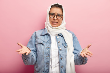 Studio shot of annoyed emotional woman wears modern Muslim clothes and white headdress, spreads hands with displeasure, has specific appearance, follows Islamic religion. People and faith concept