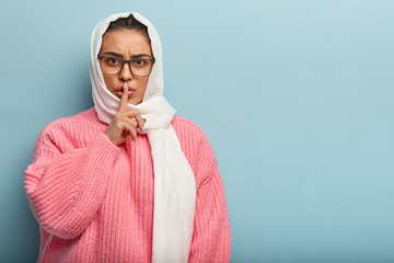 Serious angry Arabian woman holds finger on lips, says quiet please, asks for keeping silence, wears spectacles, white veil and pink jumper, isolated on blue background. Secret and ethnicity concept