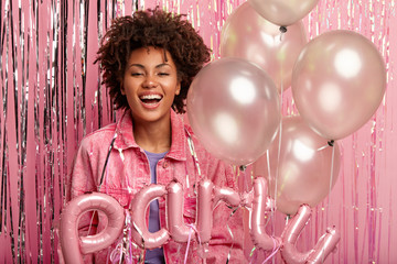 Horizontal shot of optimistic dark skinned girl smiles with pleasure, wears stylish outfit, holds party balloons and accessories, expresses positive emotions, isolated over pink studio background