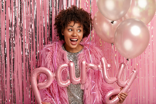 Good humored lovely dark skinned female has curly hair, focused aside dressed in long fur coat, holds air balloons glad to organize unforgettable party with peers stands over decorated pink background