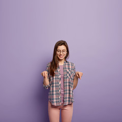 Pleased glad woman with long hair, has positive facial expression, points down with fore fingers,...
