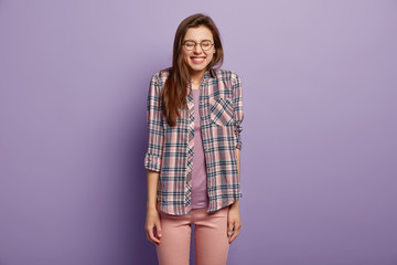 Smiling positive European woman expresses sincere emotions, hears joke or anecdote, closes eyes from pleasure, wears spectacles and checkered shirt, isolated over purple studio wall. Joy concept