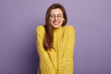 Isolated portrait of happy woman has toothy smile, closes eyes, feels pleasure from good compliment, wears glasses and yellow jumper, stands over purple wall. Positive emotions and feelings concept