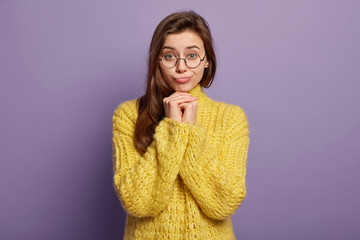 Waist up shot of pretty brunette female feels lonely and sad, keeps hands under chin, upset by bad news, wears yellow jumper, stands over purple studio wall. Human facial expressions and feelings