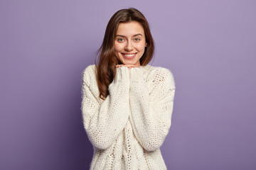 Headshot of optimistic lovely woman with straight hair, keeps hands under chin, wears white jumper, looks at camera with happiness, poses against purple background. People and emotions concept