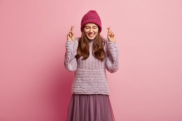 Obraz na płótnie Canvas Isolated shot of happy pleased European woman believes in luck, closes eyes in pleasure, smiles broadly, wears knitted jumper, skirt and headgear, poses over rosy wall, still hopes for good fortune
