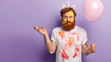 Confused discontent red haired man with bristle, raises palm and smirks face, being entertainer on holiday, wears unicorn horn and untidy white t shirt with traces of watercolours, isolated on purple © WHstudio Leushin N