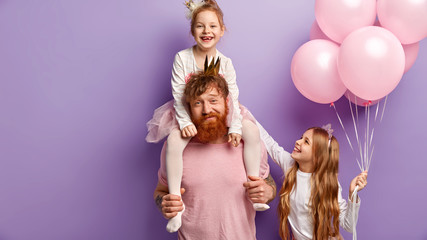 Busy redhead bearded man gives piggyback to little daughter, older sister stands near, holds air balloons, stand over purple background, celebrate holidays together. Family and celebration concept