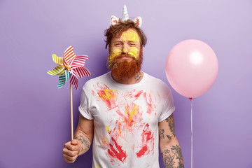 Studio shot of dissatisfied redhead man holds toy windmill and helium balloon, has face dirty with...