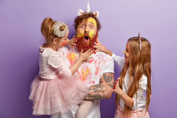 Surprised dad and two girls play together at home, paint faces with watercolours, have fun, show...