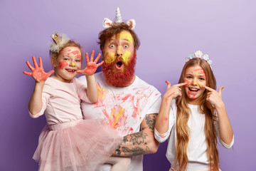 Small female child shows hands soiled with colourful gouache paintings, her glad sister has stains of watercolour on face, stunned father stares at camera, have fun before mother returns from work.